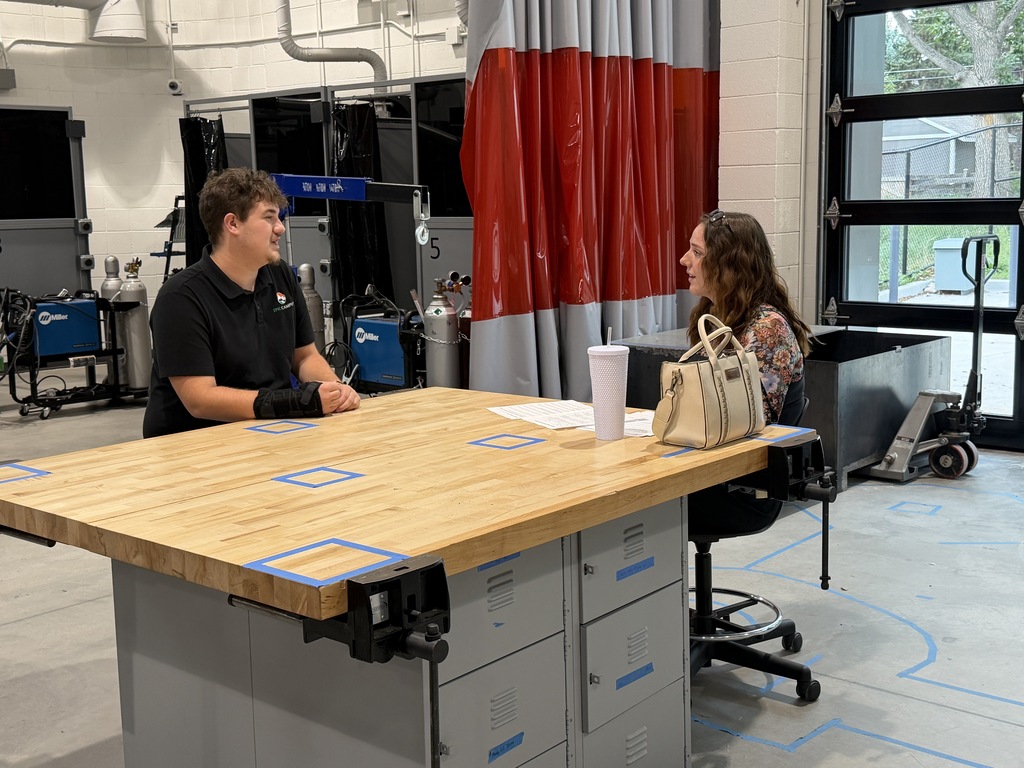 A man and a woman are seated at a wooden table in a workshop, facing each other.