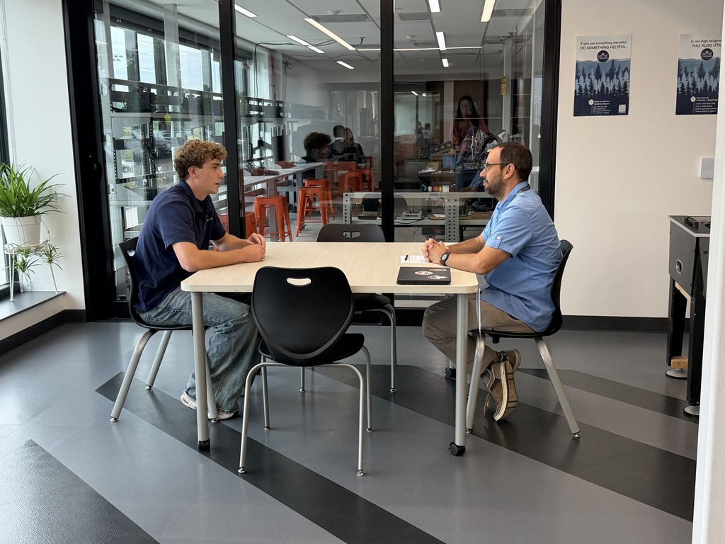 Two individuals seated at a table in an office, facing each other with a plant and glass walls in the background.