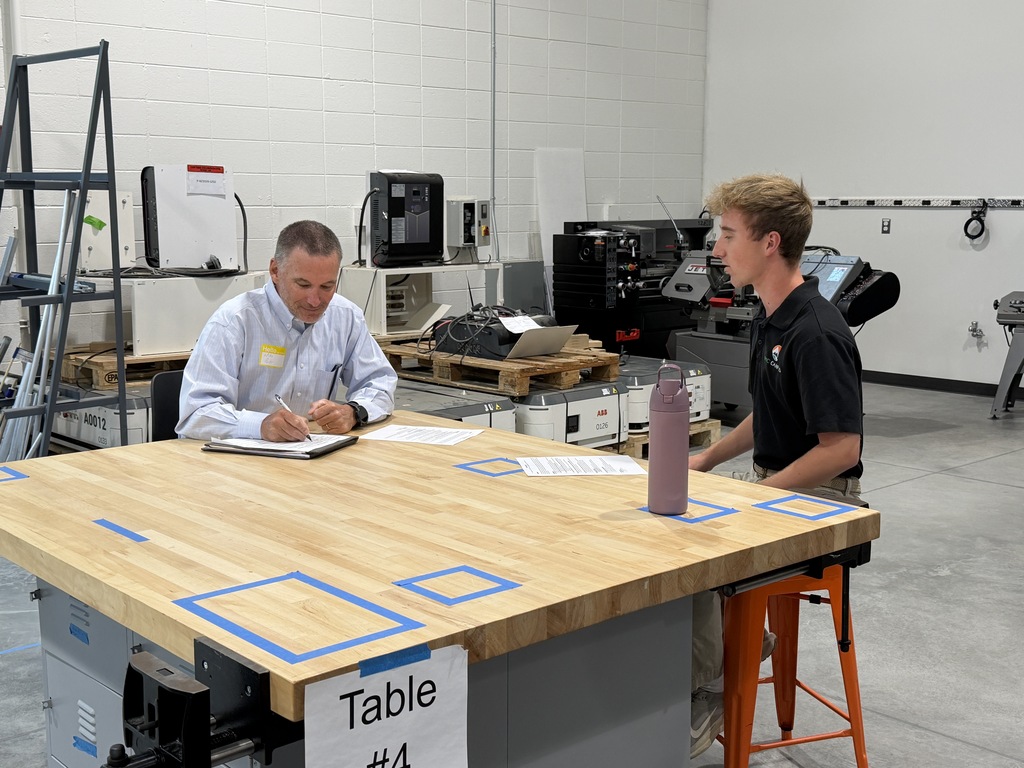 Two people seated at a wooden table in a workshop. One person writing on paper.