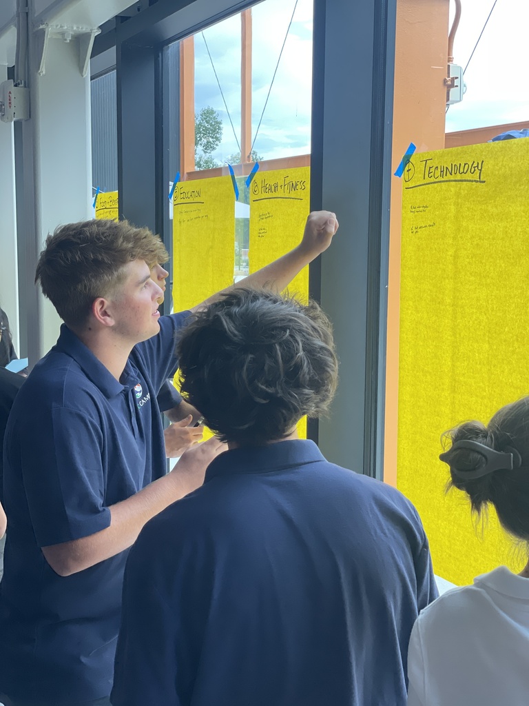 Two individuals in blue shirts examine a yellow bulletin board. A third individual with a ponytail is also looking at the board.