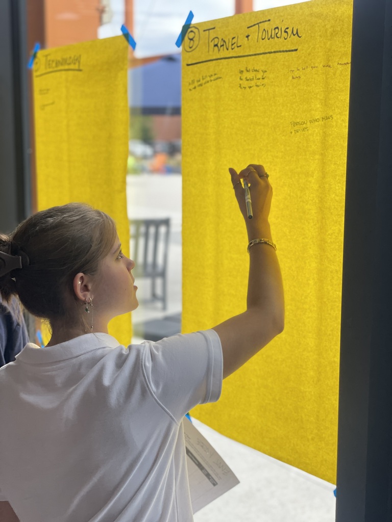 A woman writes on a large yellow poster with the words "TRAVEL & Tourism" in an indoor setting.