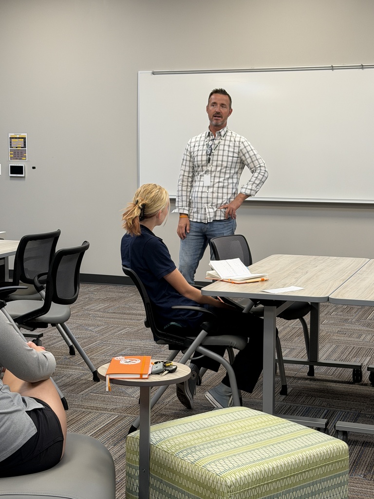 A man and woman seated in a classroom with a whiteboard and tables; the man stands and speaks.