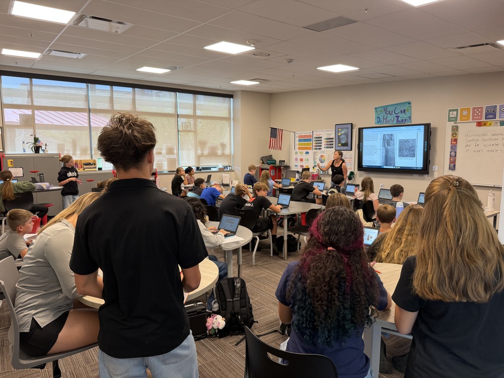 Classroom with students seated around tables with laptops. A teacher is at the front, next to a screen.