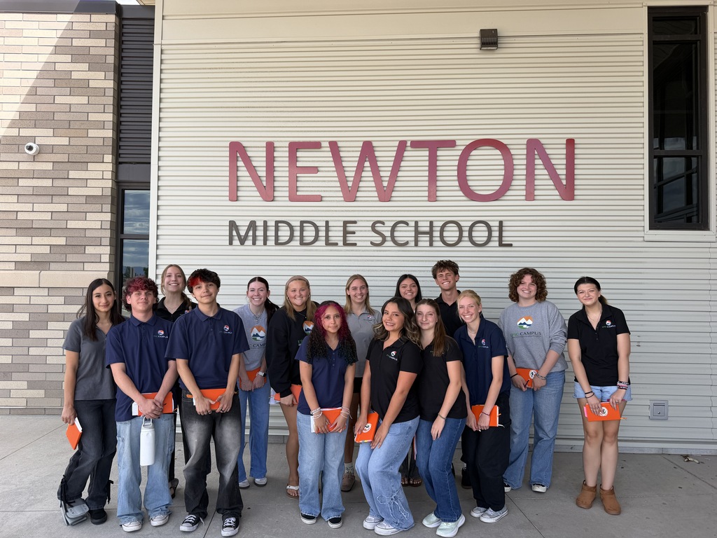 Students stand in a line in front of the Newton Middle School sign, holding orange folders.