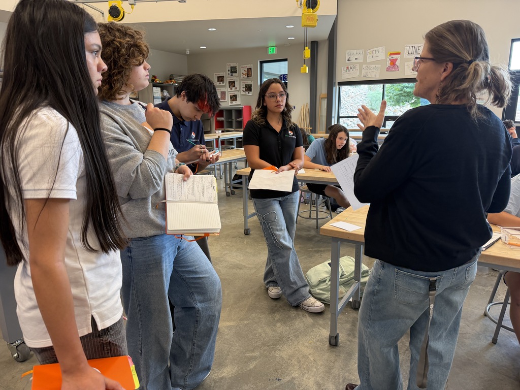 Group of students standing in a classroom with a woman in front holding papers. Bright lights on ceiling.