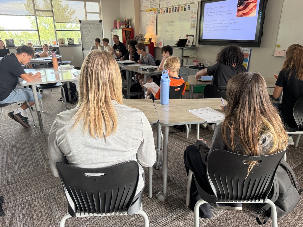 A classroom with students seated at tables. A teacher presents at the front. A monitor is mounted on the wall.