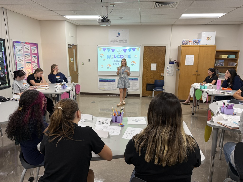 A woman speaks at a projector screen in a classroom. Students sit in rows, listening. Tables hold papers and supplies.