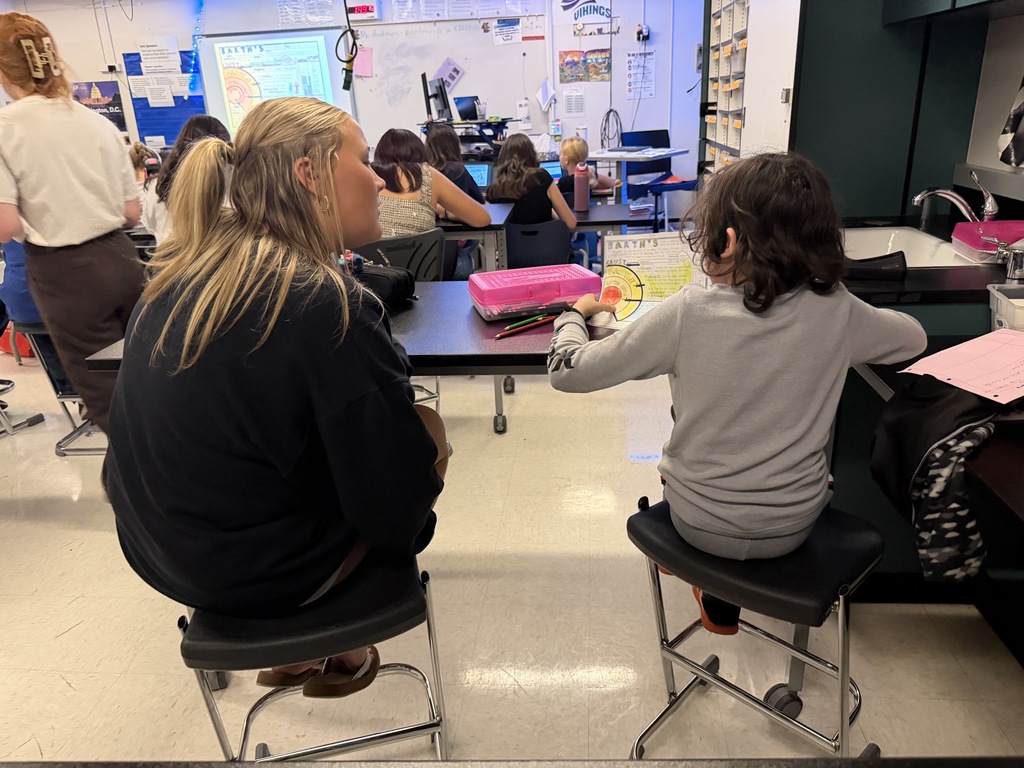 Woman sitting at table helping a child with schoolwork in a classroom with other students.