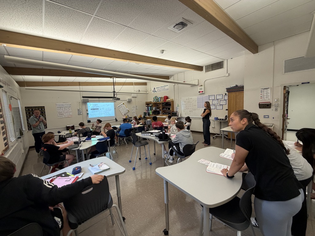 A classroom with a teacher at the front, students seated at desks, and a presentation screen.