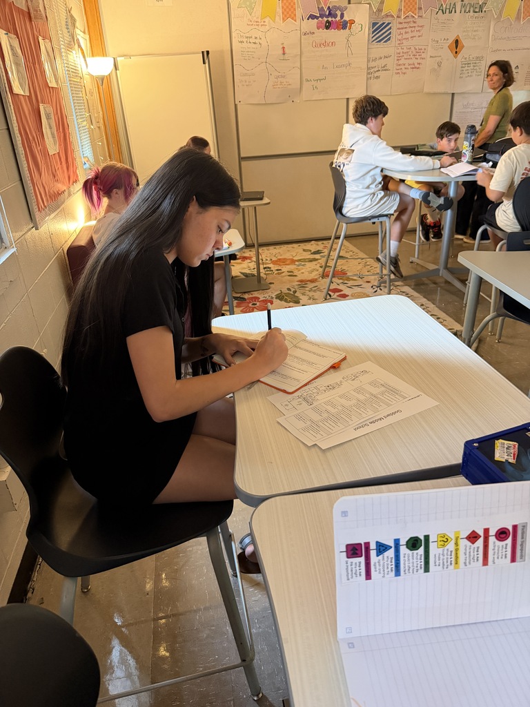 A person sits at a table writing with a pen on a paper. Others are seated around tables.