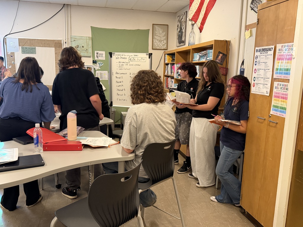 A group of people in a classroom setting, some standing, some sitting. A table with chairs, books, and a red folder is in the foreground. A whiteboard and bulletin board are in the background.
