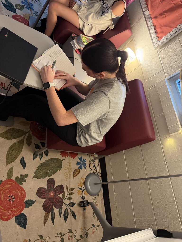 A person sits at a table writing on paper. A second person sits next to them. Bright overhead lights and a floral rug.