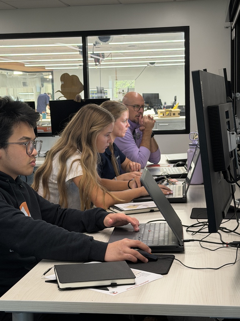 Four individuals sit around a desk in a bright room, each using a laptop. Monitors, notebooks, and pens are on the desk.