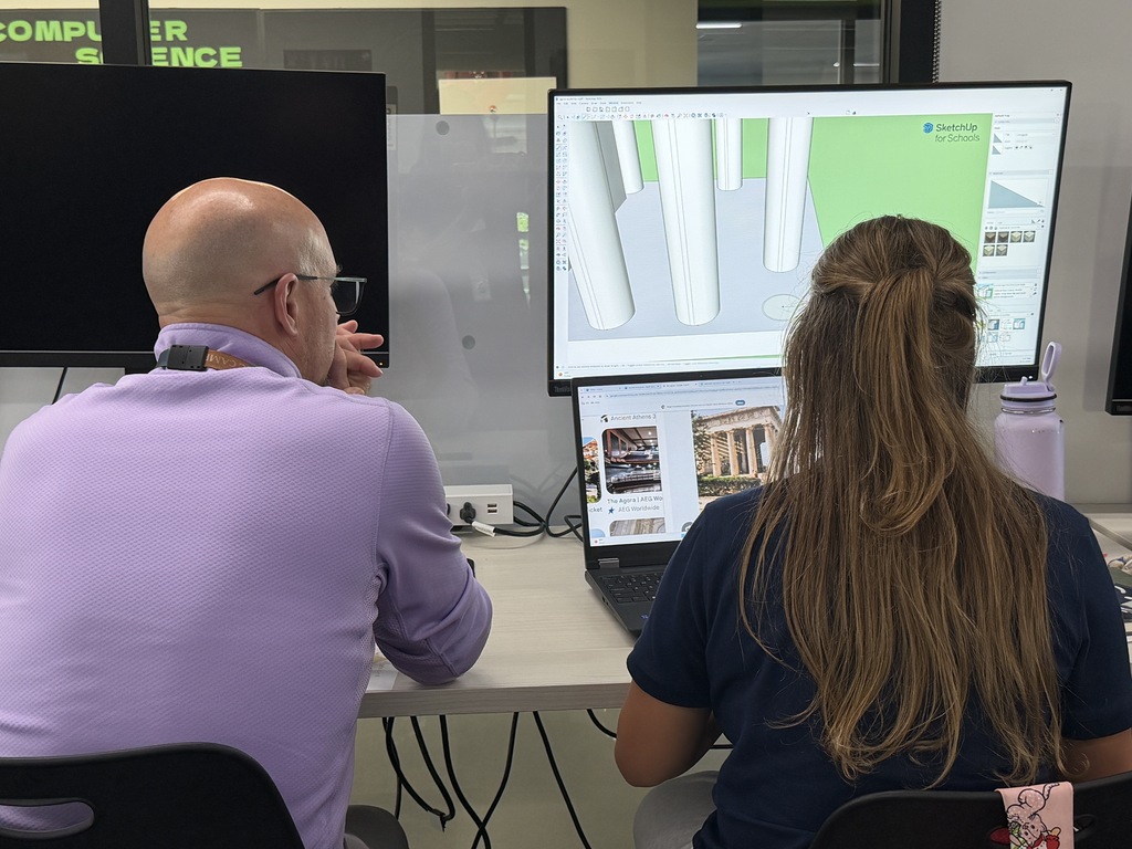A man and woman work at a desk, with monitors displaying a 3D model and a laptop.