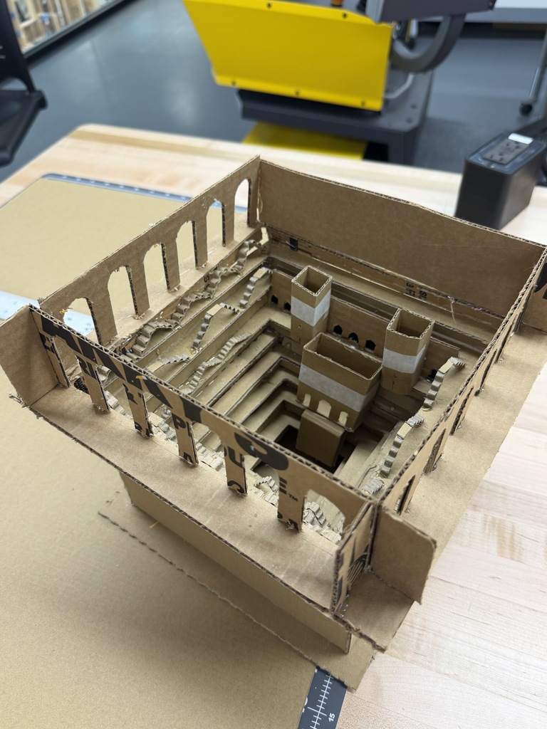 A brown cardboard model of a staircase sits on a wooden surface. A yellow machine is in the background.