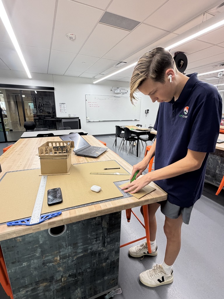 A person wearing a blue polo shirt and shorts stands at a desk cutting a piece of cardboard.