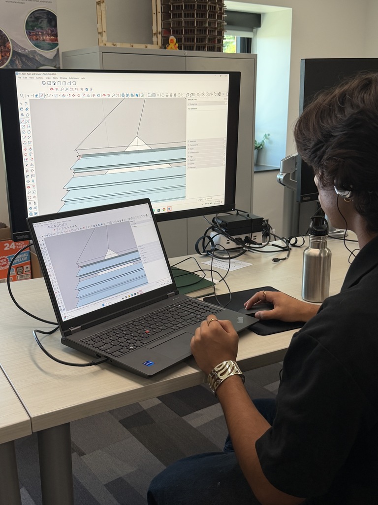 Person sitting at a desk, using a laptop and mouse. Large monitor behind the laptop shows a graph.