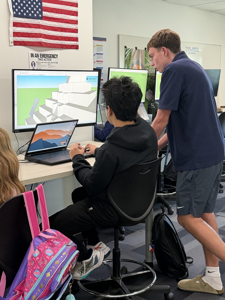 A student sits at a desk with a laptop and monitor, while a teacher walks past. An American flag is visible on the wall.