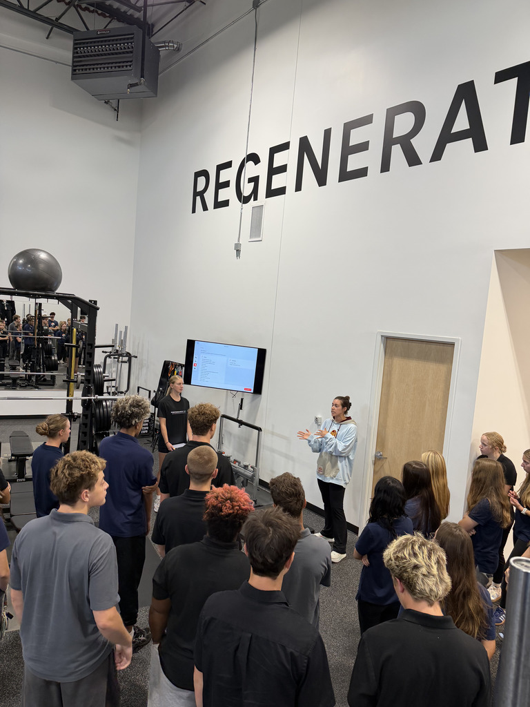 A group of people in a gym, listening to a woman speaking in front of a screen.