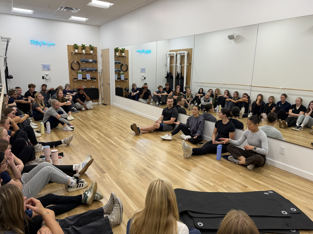 A group of people seated on a floor, some standing, in a studio with wooden floors and mirrors.