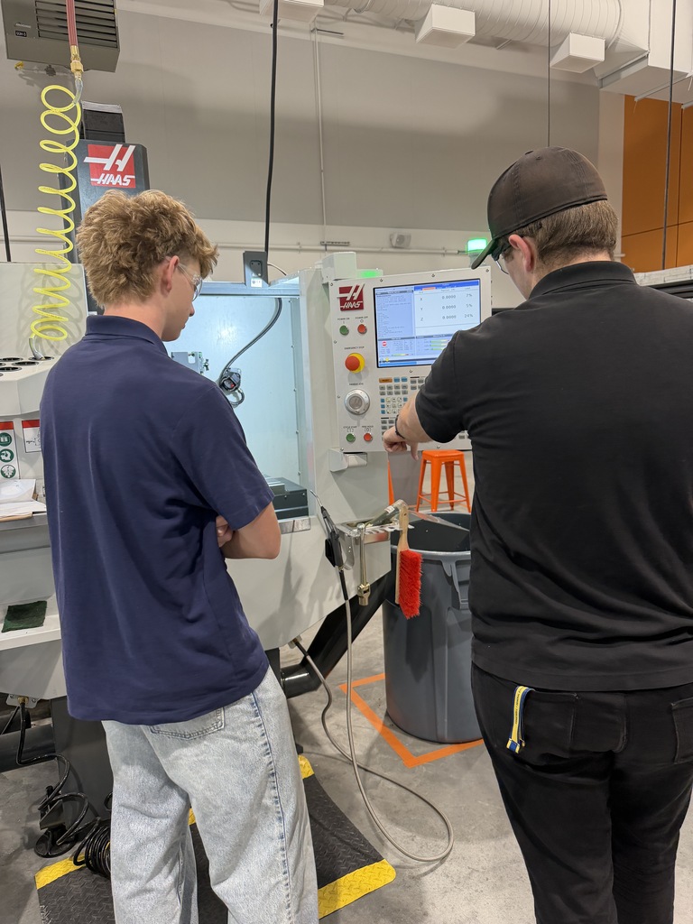 Two men in a factory, one standing and the other adjusting a machine, with a trash bin nearby.