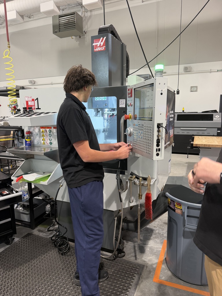 A person stands before a large industrial machine in a factory, adjusting a control. Another individual stands nearby.