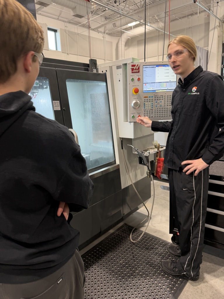 Two individuals in a factory. One man is gesturing to a large machine with a screen.