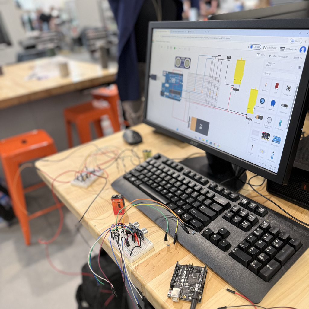 A workspace with a monitor, keyboard, wires, and circuit boards on a desk. A person in the background.