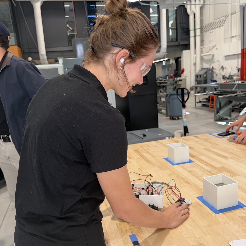 Woman working with electronic equipment on a table. Behind her, a man stands near a machine.