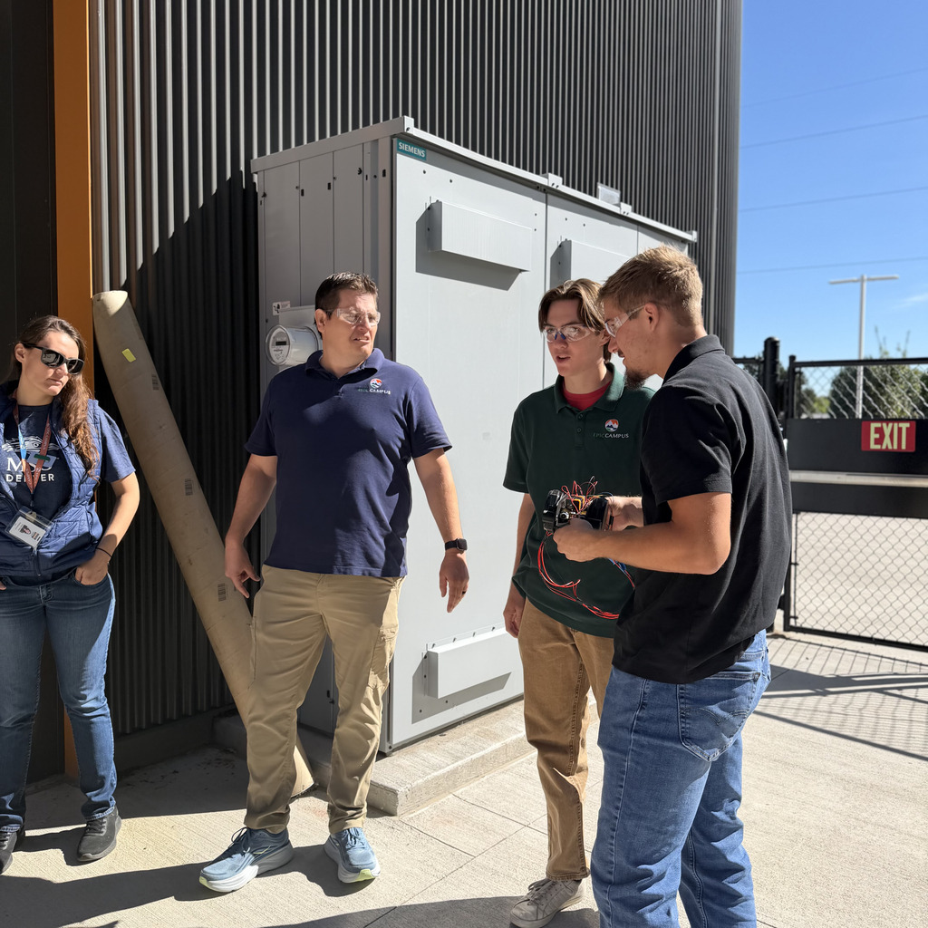 Four people stand in front of a building. One person holds a tool. They look at a large gray box with a handle.