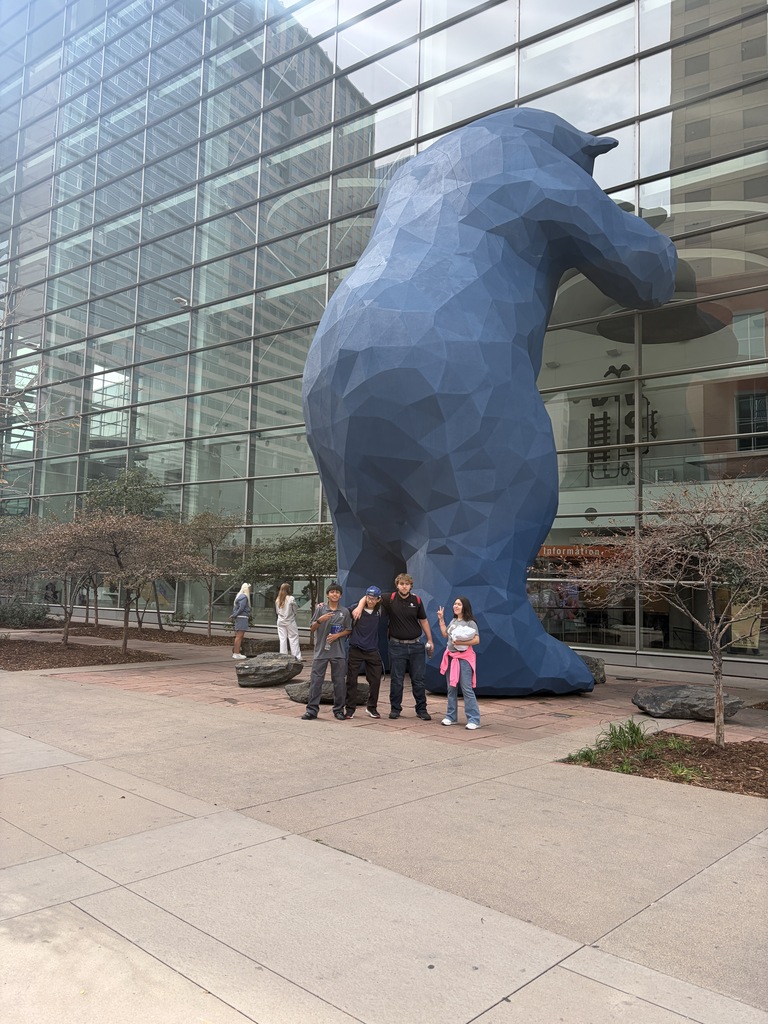 Group of people standing in front of a large blue bear statue, outside a building with glass walls.