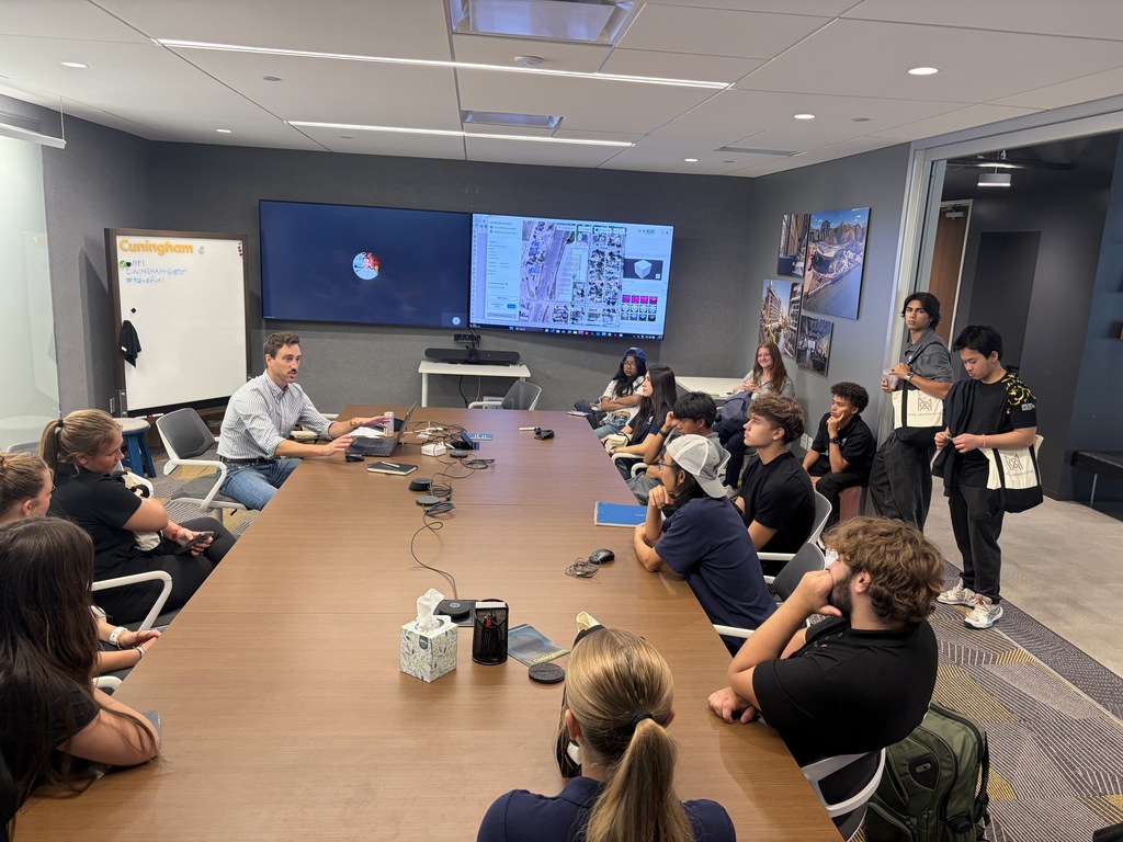 A group of people seated around a long table in a conference room with monitors and a whiteboard.