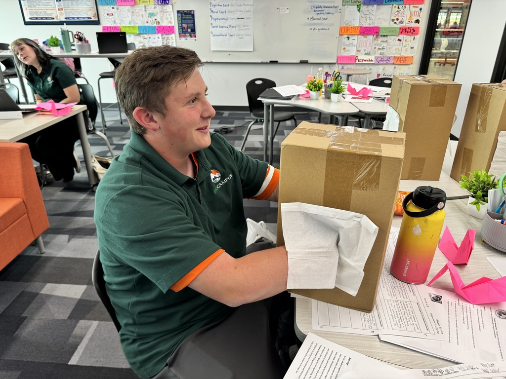 A person wearing a green polo sits at a desk, handling a box while wearing gloves.