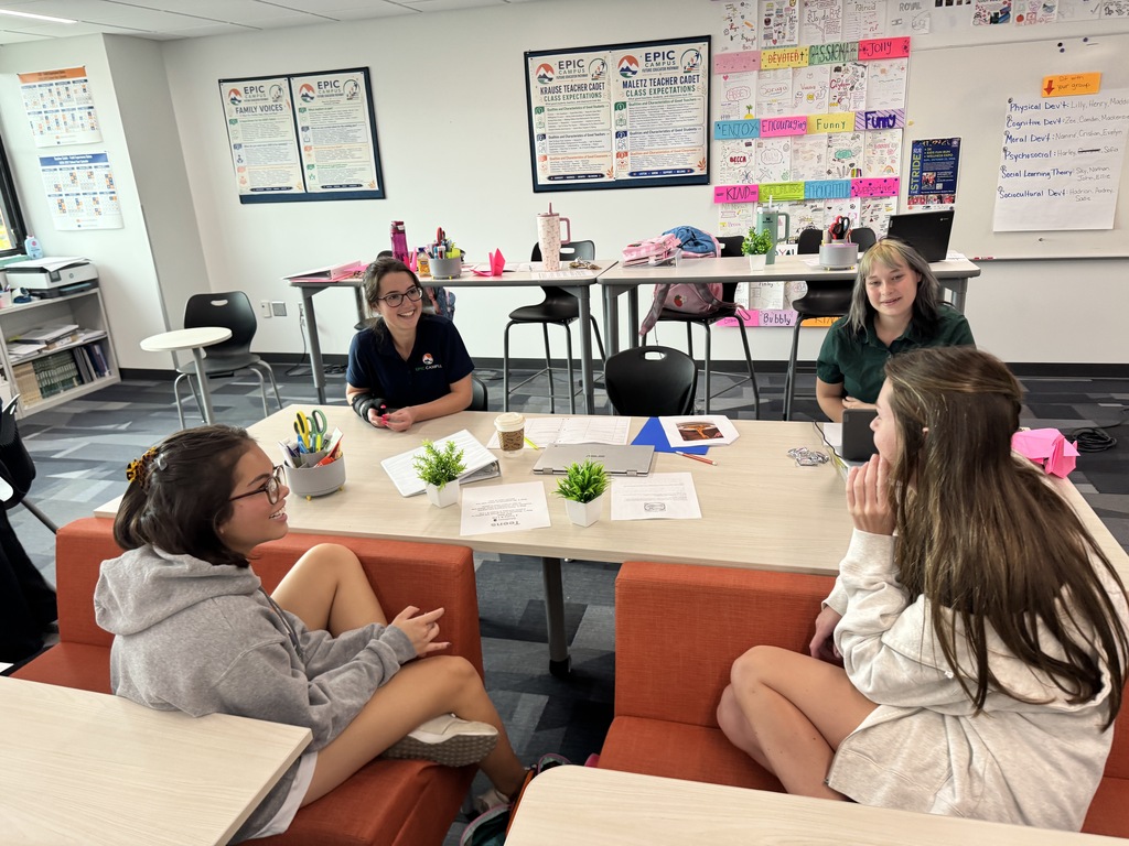 Four individuals are seated at a table, engaged in conversation. Three are women, one with glasses. The setting is a room with desks, chairs, and a bulletin board.