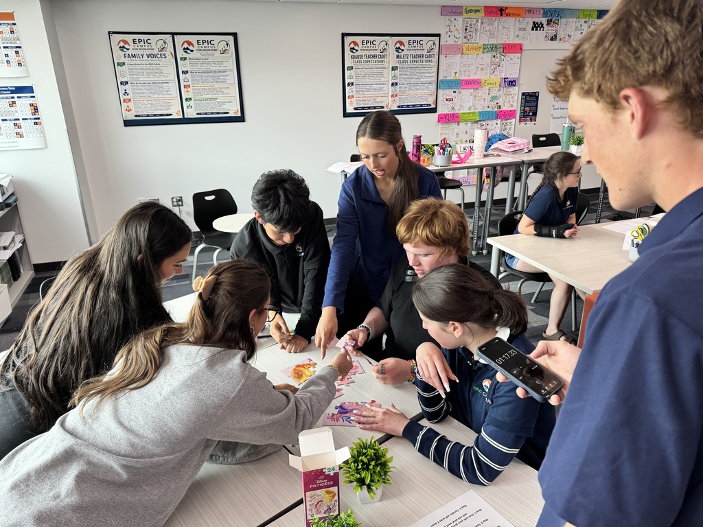 A group of people sit at tables inside a classroom. Some engage in activities. One person holds a cellphone.