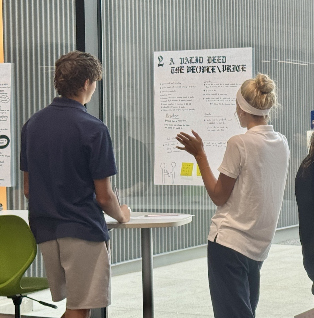 Two people in front of a table, a woman gesturing to a poster, and a man looking at it.
