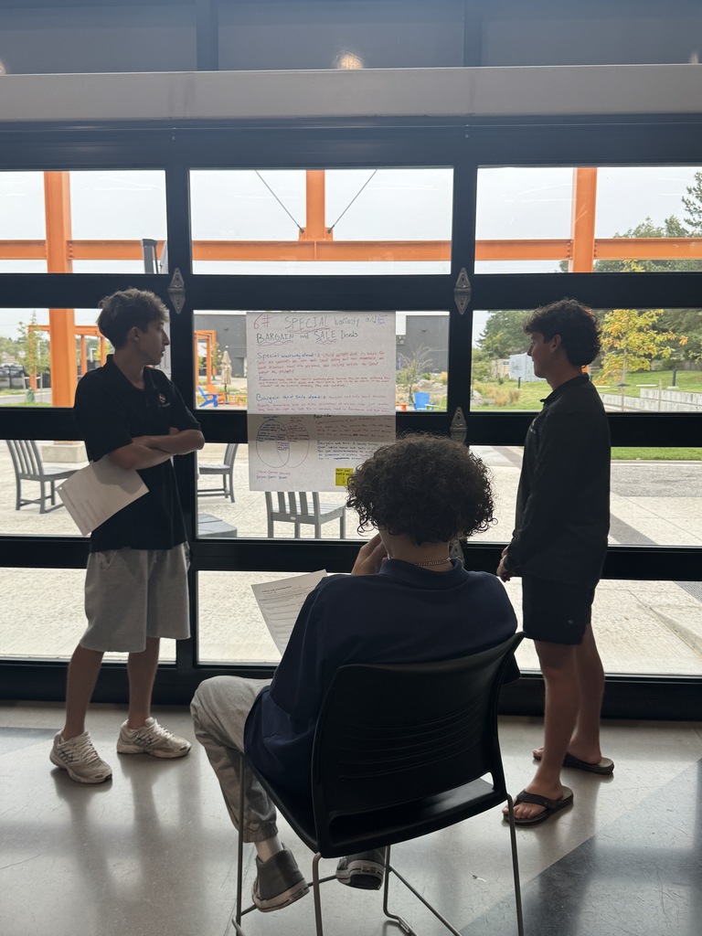 Three people stand by a large window. One sits on a chair. Two hold papers, and one looks at a whiteboard.
