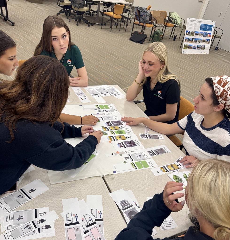 Five young women seated at a table surrounded by papers, cards, and drawings. Two of them wear green shirts.