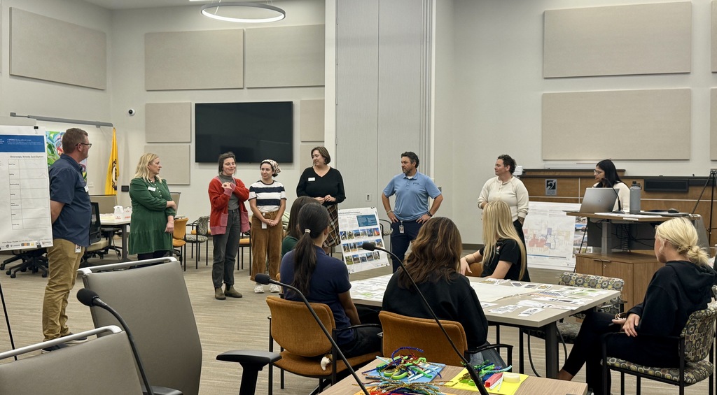A group of people stand and sit around tables in a room, some discussing. A screen hangs on a wall.