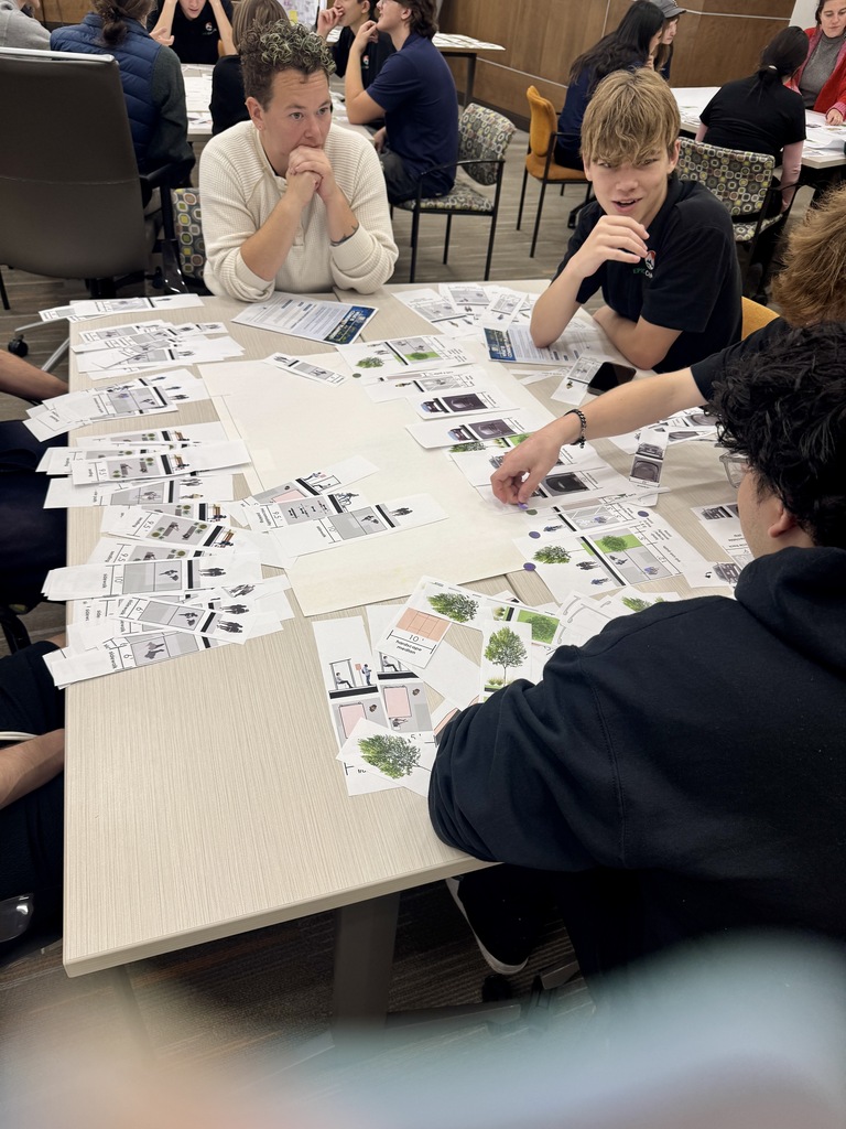A group of students seated at a table with architectural drawings spread out, some actively discussing.