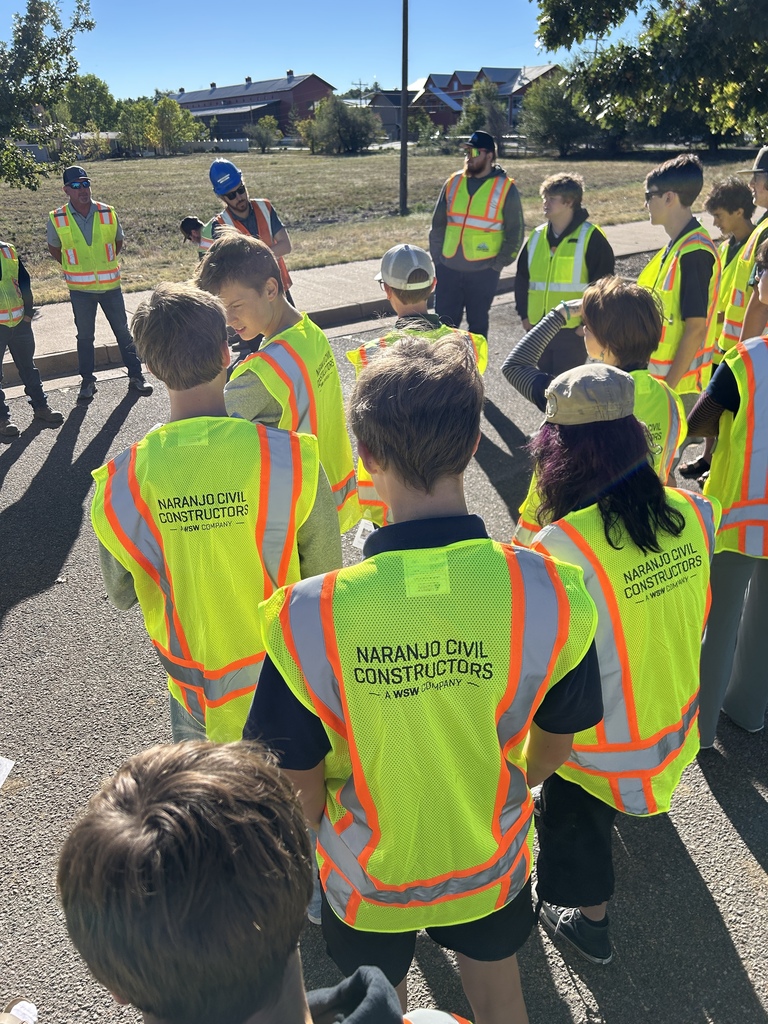 Children in yellow vests stand in a line. A grassy area is in the background with buildings.
