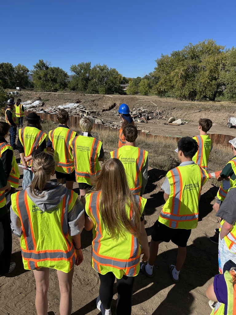 A group of people in high-visibility vests, standing on a dirt field with a blue sky.
