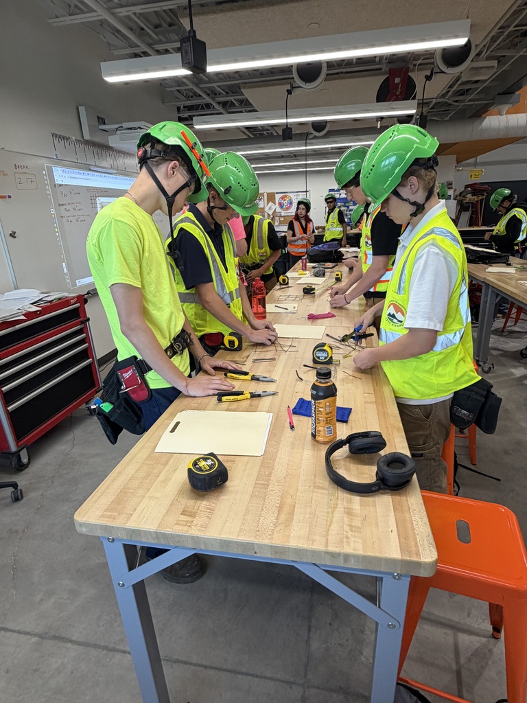 Several individuals wearing yellow vests and green helmets are engaged in a workshop. They are seated at a table with tools.