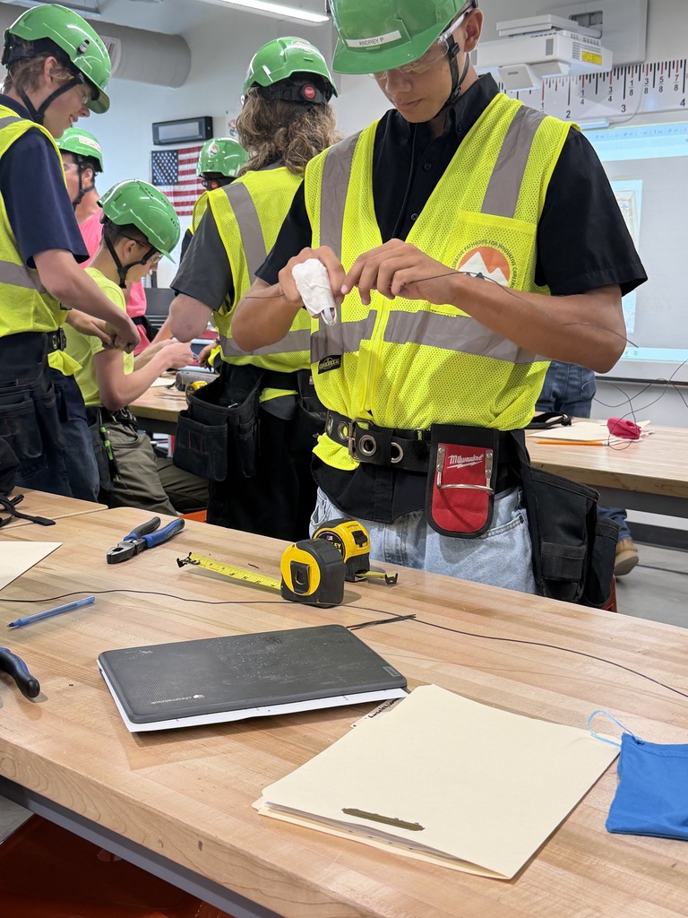 A group of people in green helmets and safety vests work on a wooden table with tools.