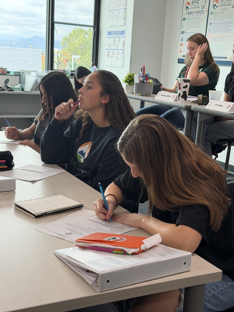 Students sitting at a table, some writing with pens, and one blowing bubbles. A bright room with glass windows.