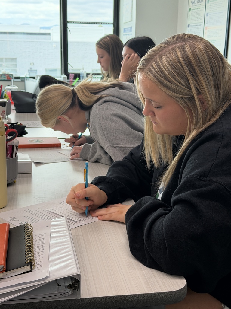Four women in a classroom setting, seated at a table, engaged in writing activities. Papers, notebooks, and pens are on the table. A window is visible in the background.
