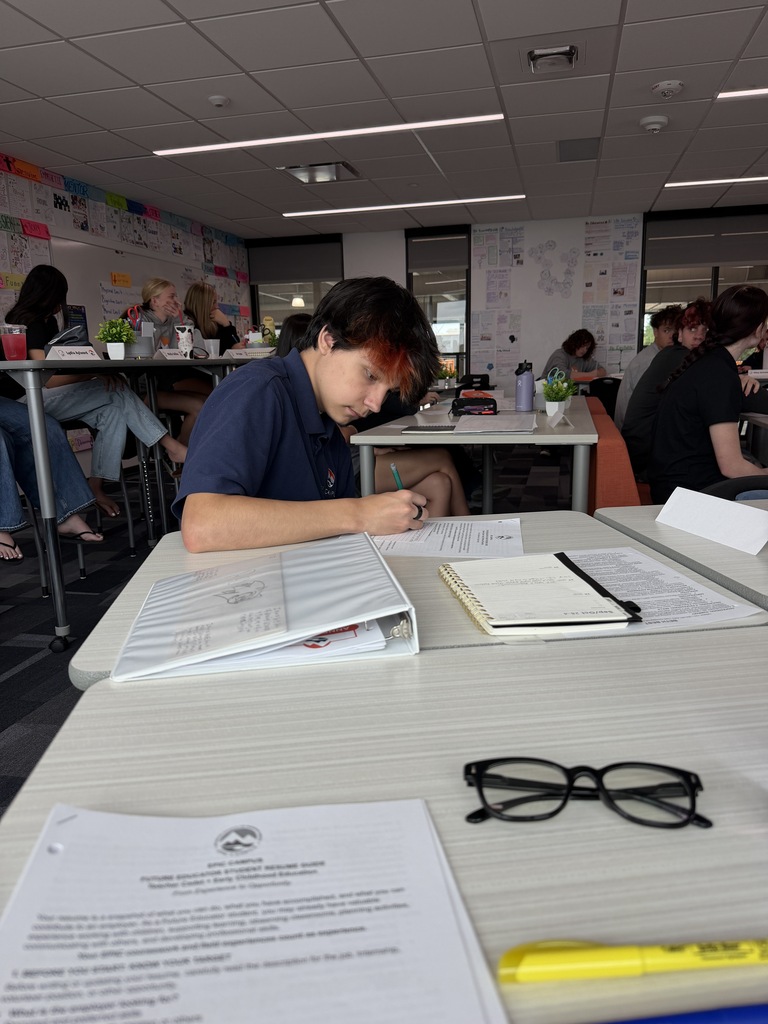A person in a blue shirt writing at a desk. Open binder, papers, and glasses on table.