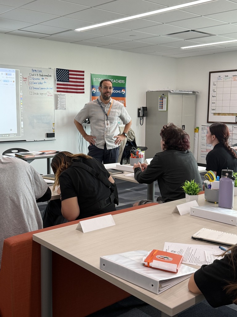 A classroom with several students seated at tables. One man stands at a whiteboard, addressing the class.