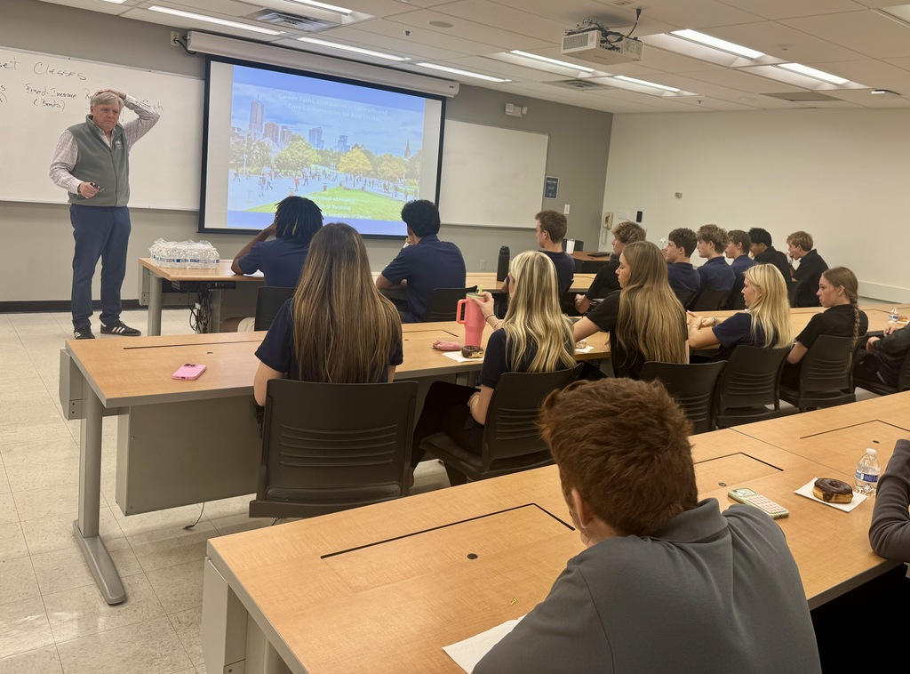 A classroom with a teacher standing by a projector screen, while students sit at desks.