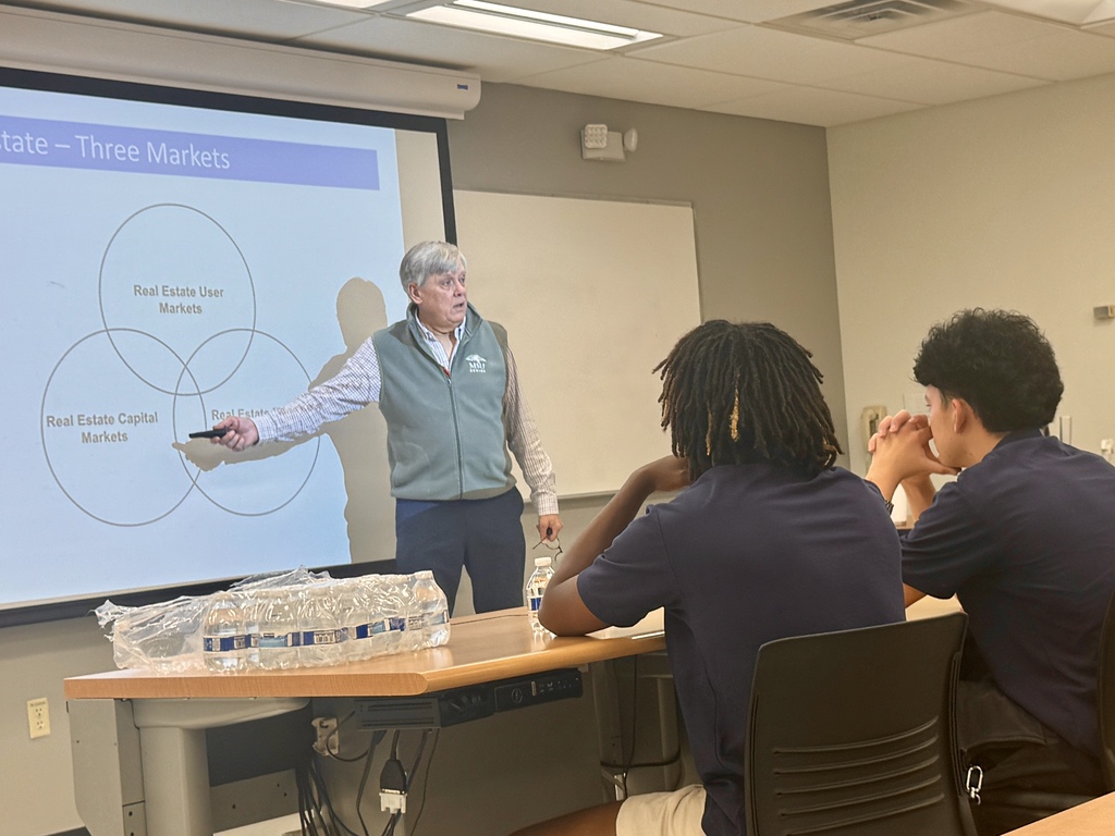 A man presents to students at a table. The person speaks with a remote in hand.